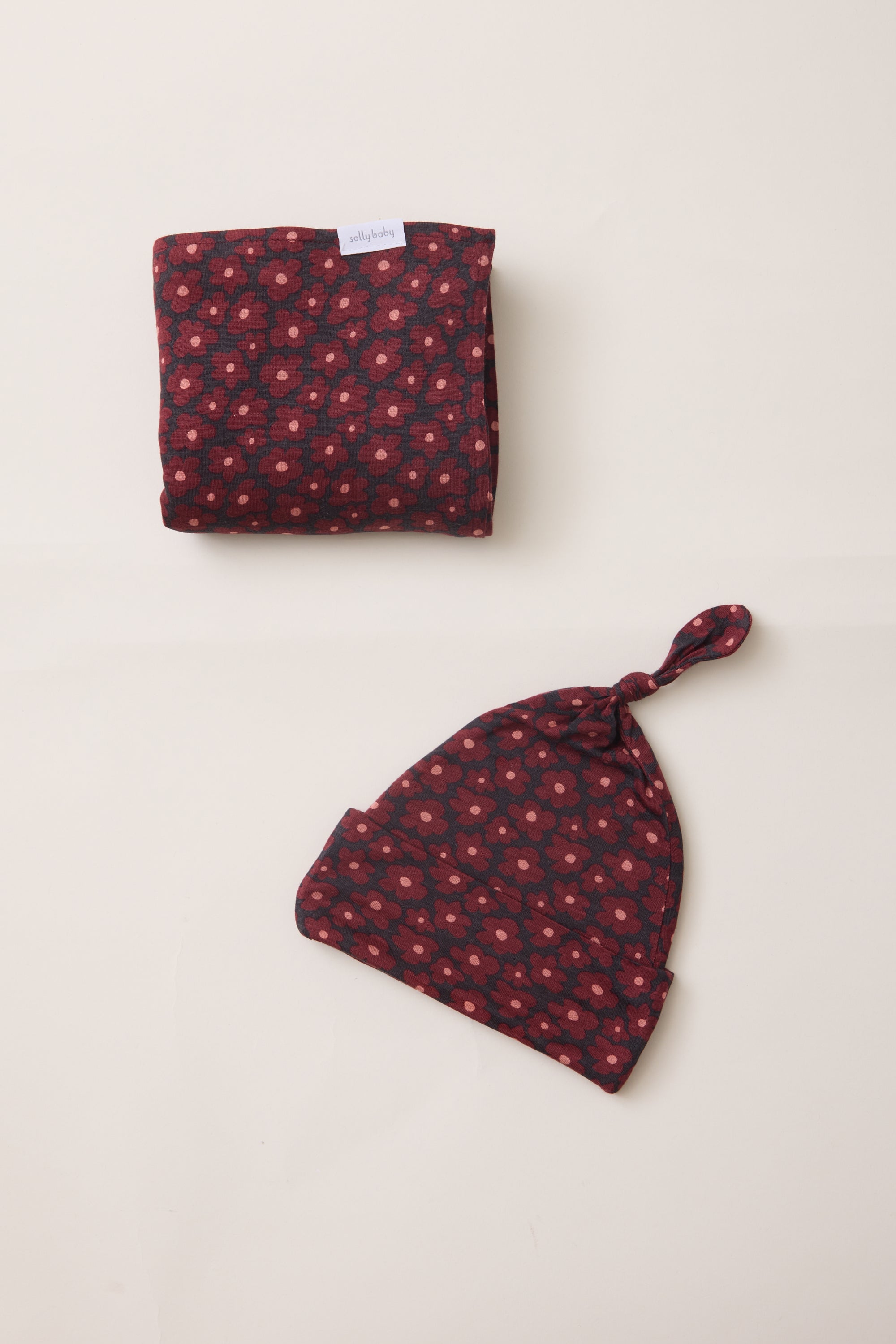 A folded matching baby set consisting of a soft cloth wrap and a knotted beanie hat, both featuring a dark red and navy blue floral pattern, laid out on a light beige background.
