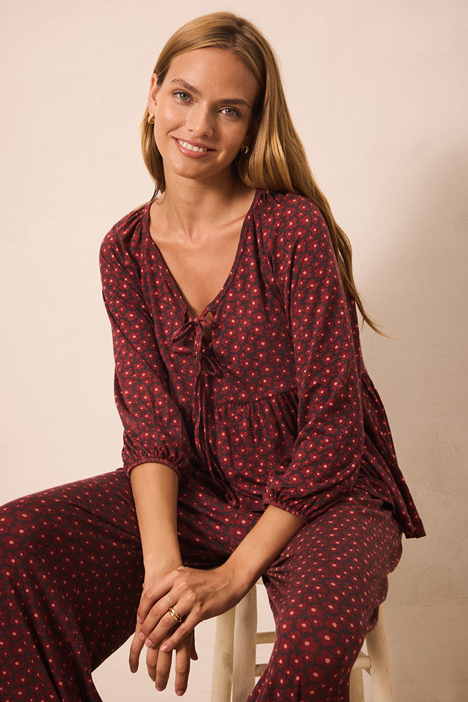 A woman wearing a matching burgundy and red floral print lounge set, sitting on a white stool against a warm beige background.