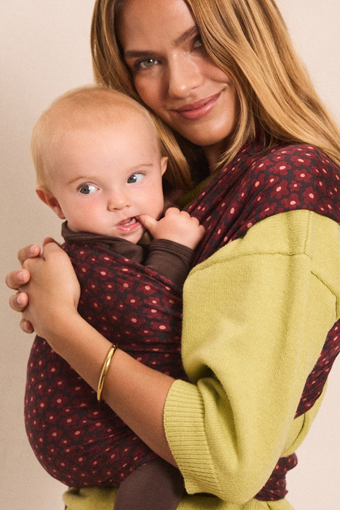 A woman wearing a green and burgundy polka-dot baby wrap carrier holds a baby against her chest. The baby is also wearing a matching brown wrap.