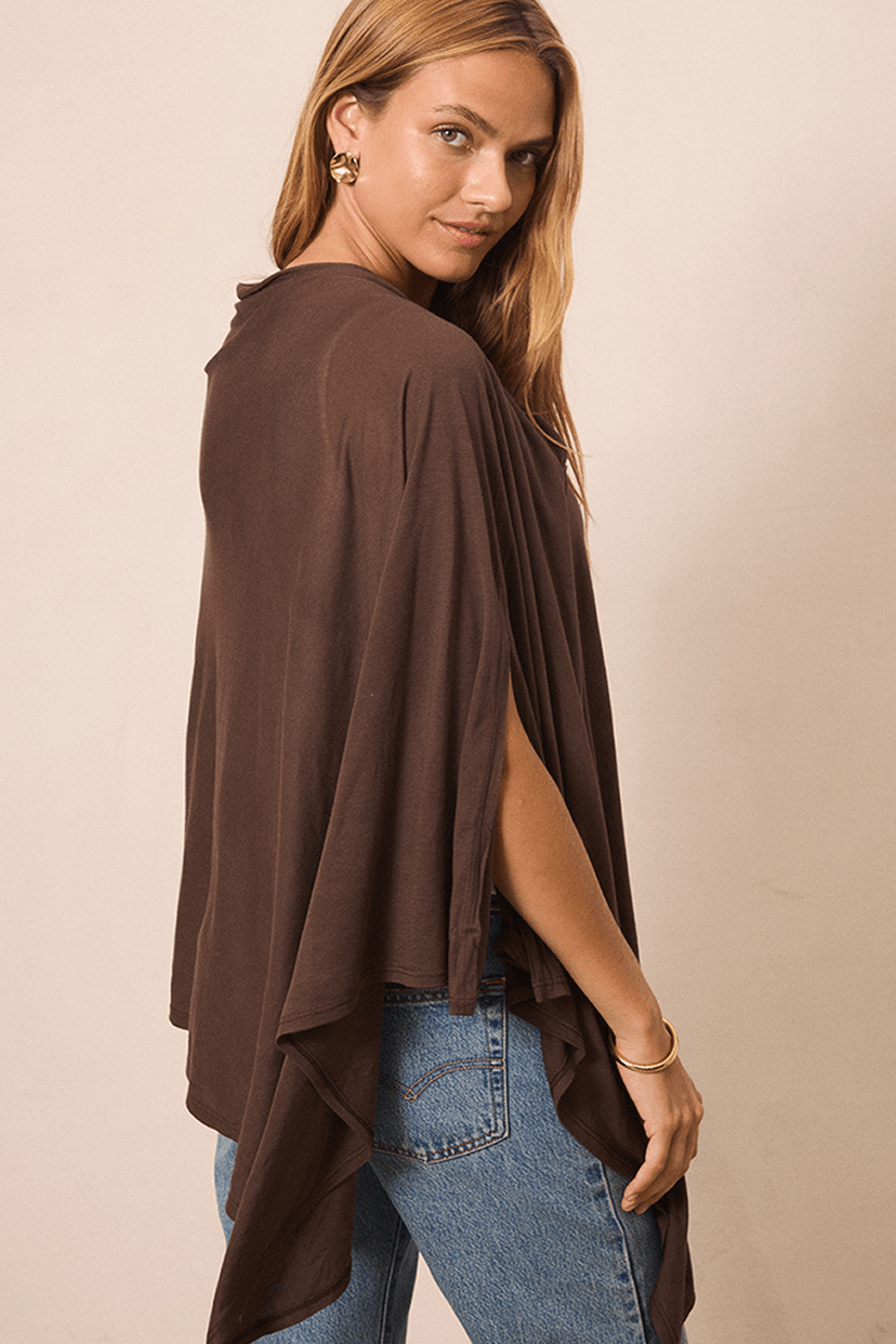 A woman wearing a brown draped poncho-style top with side slits and blue jeans, photographed against a plain beige studio background.
