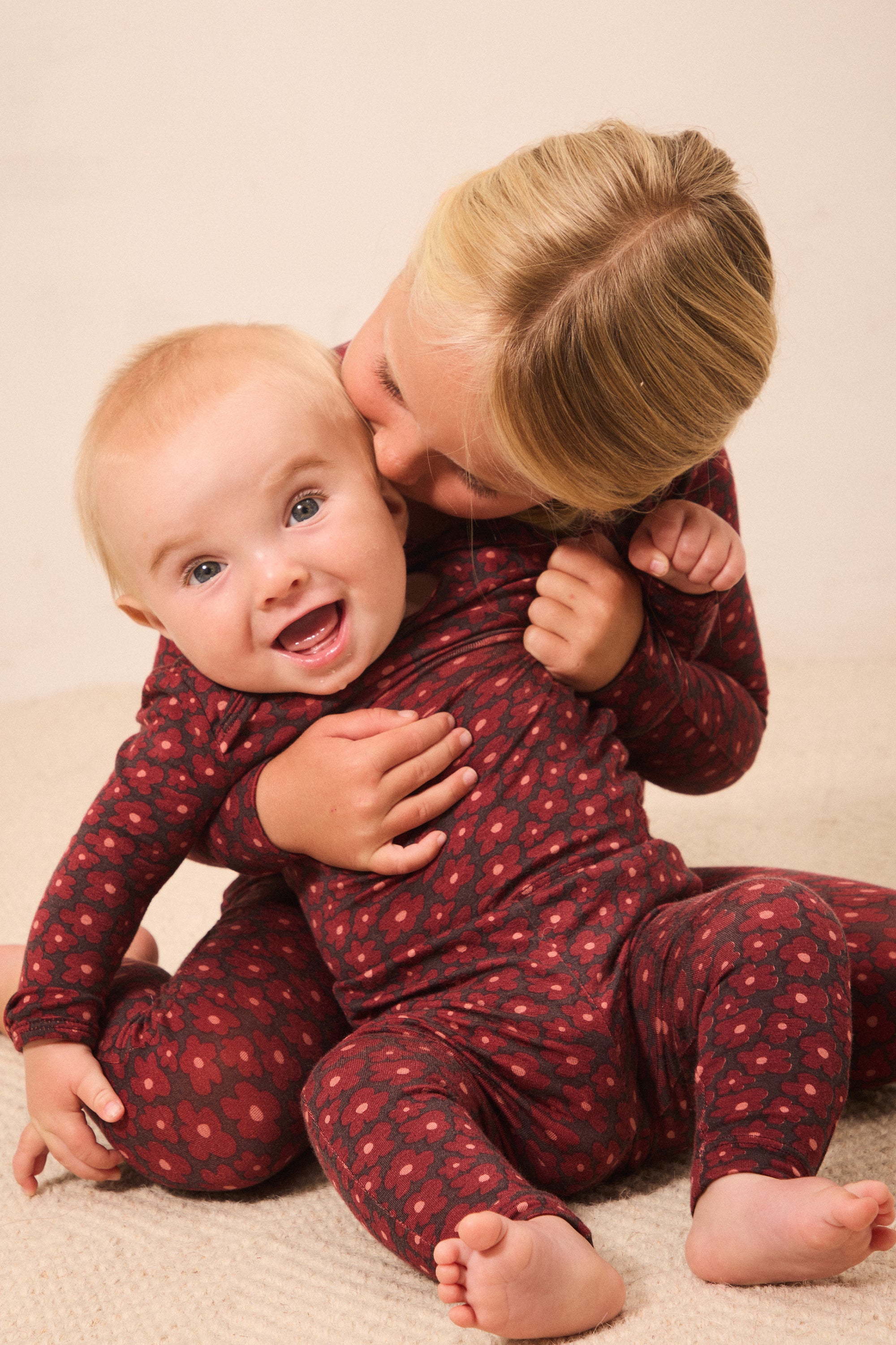 Two babies wearing matching dark red and black patterned long-sleeve footless onesies, with one baby hugging and kissing the other on the cheek, on a beige carpeted floor.