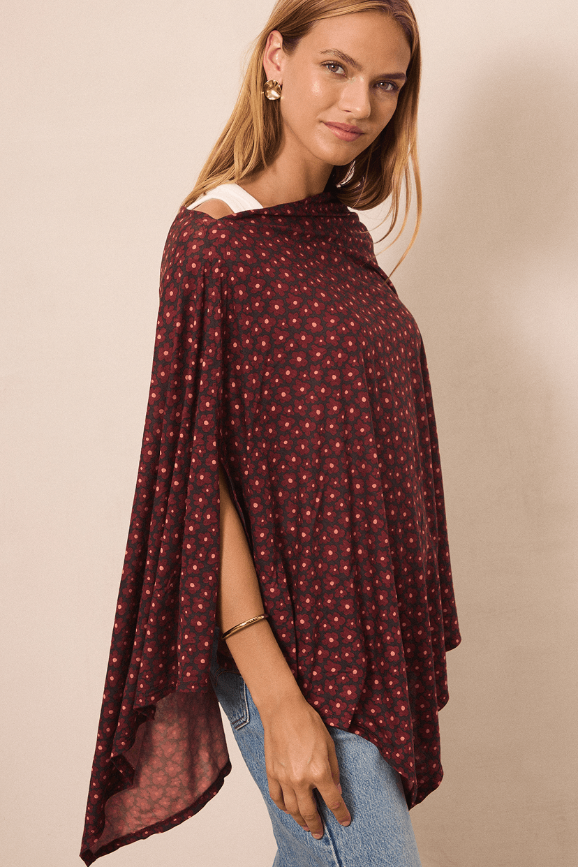 A woman wearing a dark pink and flower patterned poncho top with blue jeans, photographed against a plain beige studio background.