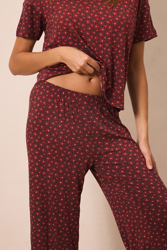 A woman wearing a matching short-sleeve top and pants pajama set in dark red with a small white floral pattern, shown from the chest down against a plain beige background.