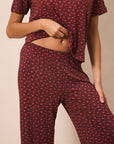 A woman wearing a matching short-sleeve top and pants pajama set in dark red with a small white floral pattern, shown from the chest down against a plain beige background.