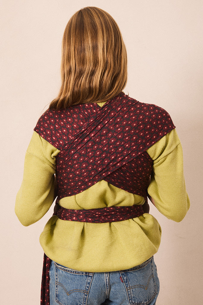 A woman viewed from behind wearing a wrap-style baby carrier or shoulder wrap in dark red with a small repeating floral pattern, worn over a yellow-green top and blue jeans against a plain beige studio background.