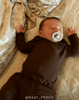 A sleeping newborn baby wearing a dark brown long-sleeve romper and tights, lying on a beige quilted bed with a patterned gold and beige blanket. The baby has a white pacifier in their mouth, and the handle of the photo is signed @NAAT_PROCH.