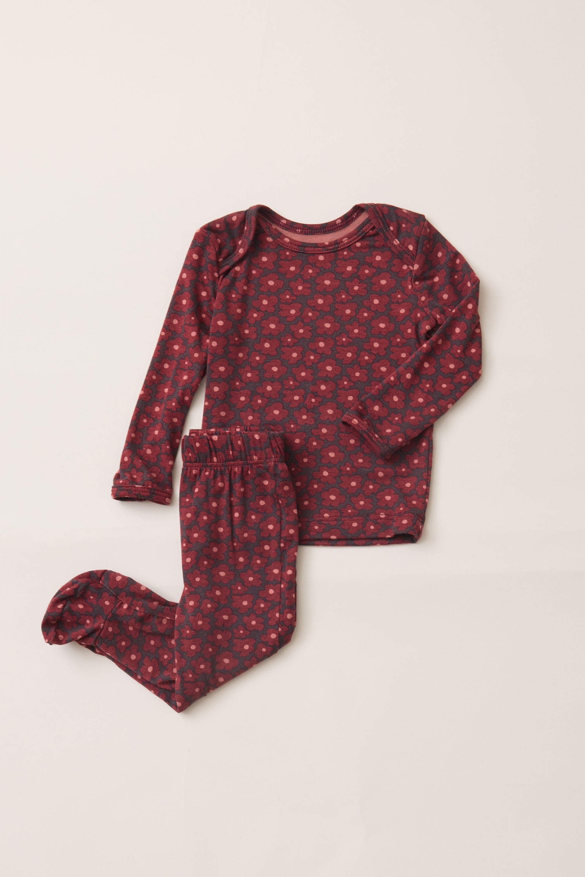 A two-piece baby pajama set featuring a long-sleeve top and footed pants in a dark red or burgundy fabric with a small pink floral pattern, laid flat on a light beige background.