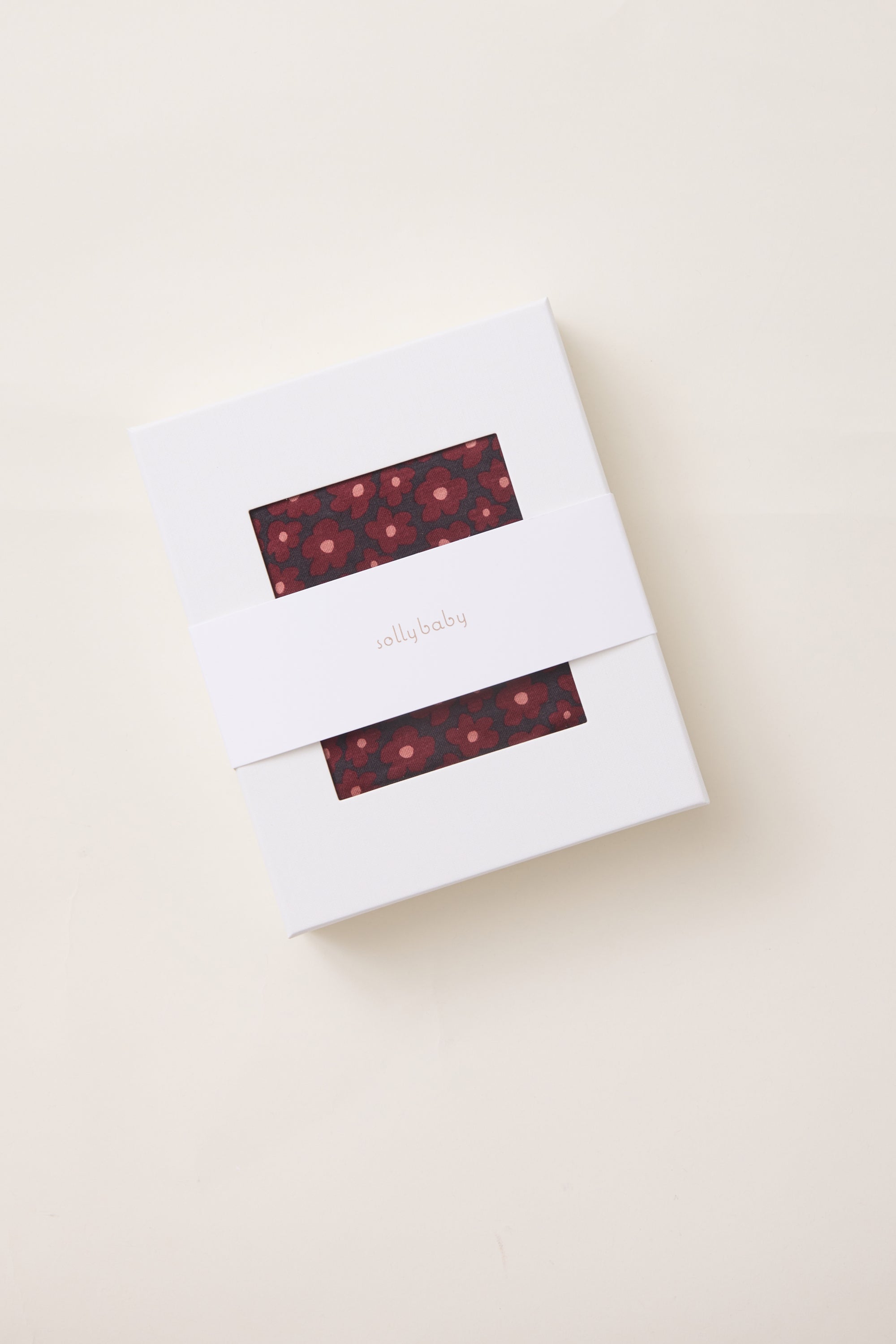 A white rectangular gift box with two horizontal cutouts showing a red floral patterned fabric, wrapped with a white paper band labeled 'solly baby' on a light beige background.