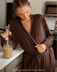 A woman in a brown wrap robe leans on a kitchen counter with a glass of iced coffee, wearing gold hoop earrings and a ring. The text '@HEALINGWITHHANNA' is visible at the bottom.
