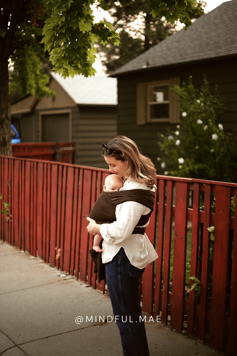 A woman wearing a white shirt and dark jeans carries a baby in a brown wrap carrier outdoors on a sidewalk next to a red wooden fence.