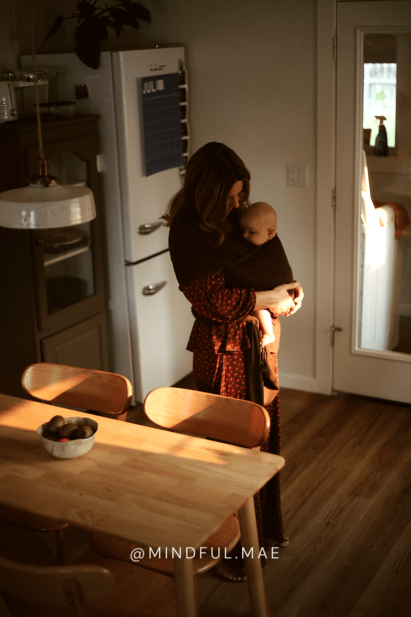 A woman wearing a dark brown and red patterned wrap dress holds a baby in a brown baby wrap carrier inside a warmly lit home kitchen.