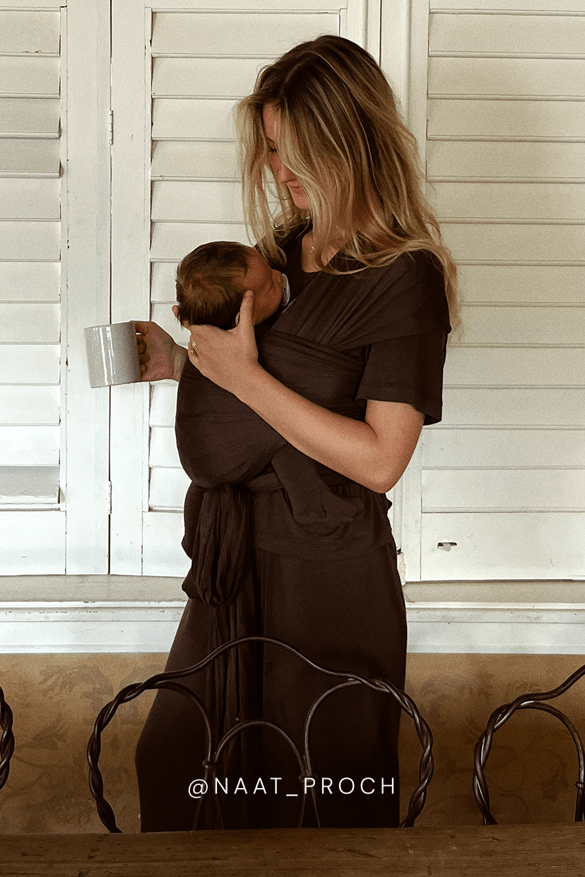 A woman wearing a dark brown baby wrap dress holds a baby against her chest. The baby wrap dress is a short-sleeve wrap style with a tie waist.
