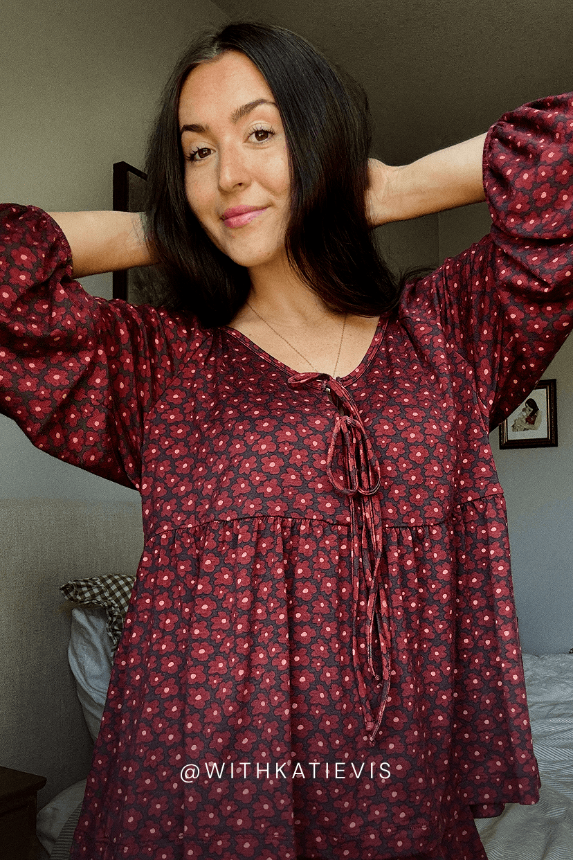 A woman with dark hair wearing a dark pink flower patterned long-sleeve top with a small pink floral pattern and a tie-neck detail, posing with her arms raised in a bedroom setting.