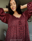 A woman with dark hair wearing a dark pink flower patterned long-sleeve top with a small pink floral pattern and a tie-neck detail, posing with her arms raised in a bedroom setting.