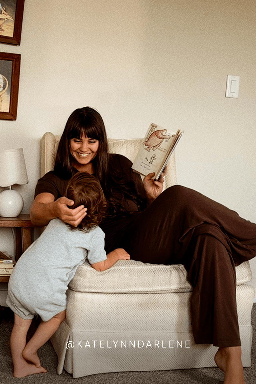 A woman in a dark brown outfit relaxes in a cream armchair, reading a book to a young child who is leaning against the armchair. The scene is set in a cozy, neutral-toned room with a side table and lamp visible.
