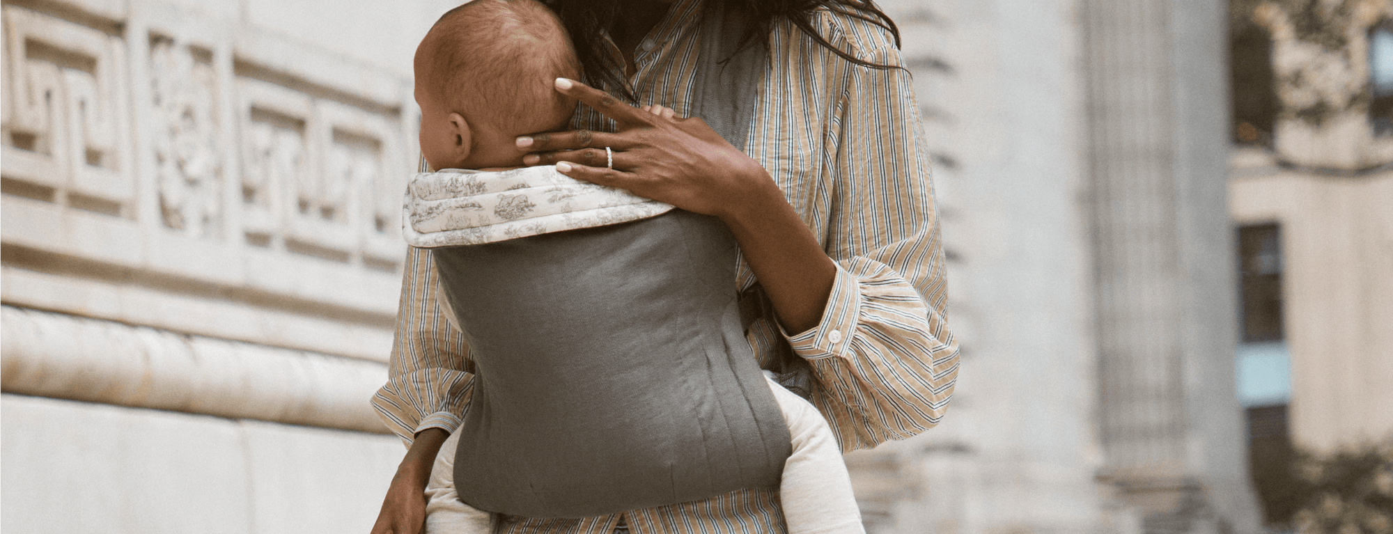Mother wearing a neutral-toned baby carrier with baby facing inward, featuring a soft toile trim.