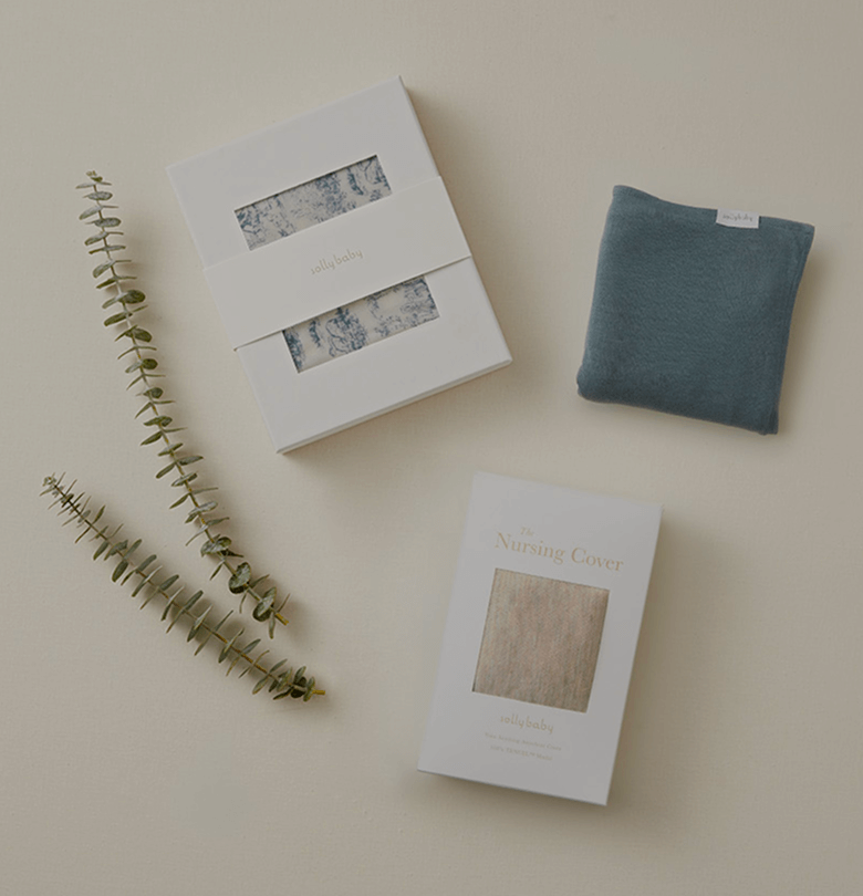 Flat lay of a beautifully curated gift set featuring Solly Baby essentials. A boxed set of our signature soft wrap in a delicate blue toile design rests beside a cozy, folded fabric piece in a rich, moody blue. A thoughtfully designed nursing cover, wrapped in elegant white packaging with a soft neutral fabric swatch, completes the trio. Eucalyptus sprigs add an organic touch, all styled against a warm, neutral backdrop for a serene and timeless presentation.