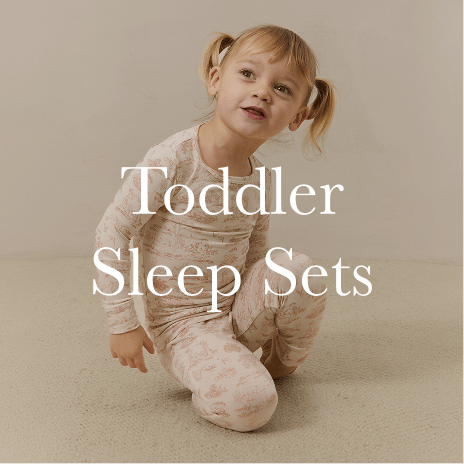 A toddler in a soft, neutral toile sleep set sits on a cozy surface, smiling with an effortless sense of comfort and ease. Shop Toddler Sleep Sets.