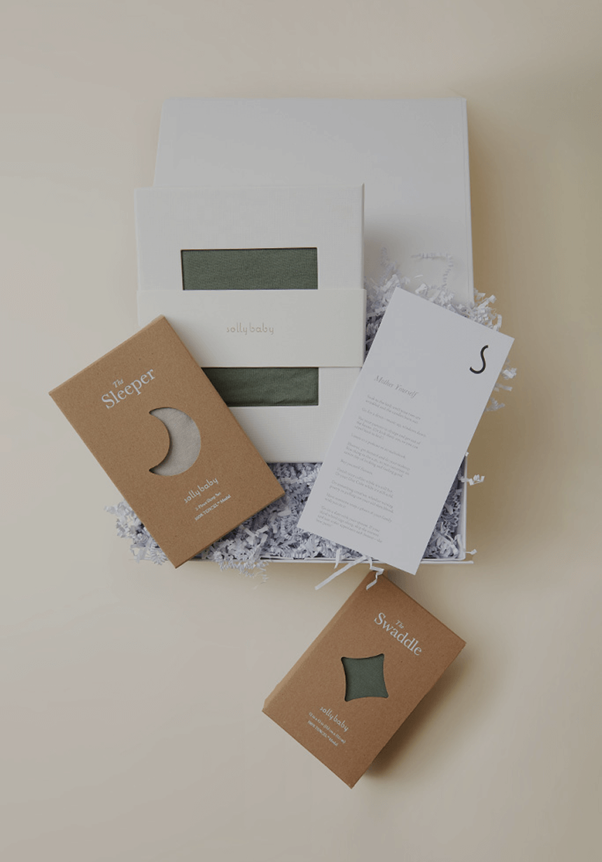 A beautifully curated Solly Baby gift set sits atop soft crinkle paper, featuring an organic cotton sleeper and swaddle in a muted sage green. Packaged in elegant kraft boxes, the set is thoughtfully designed for new parents, making it the perfect baby shower gift. A handwritten note and branded details complete the timeless presentation.