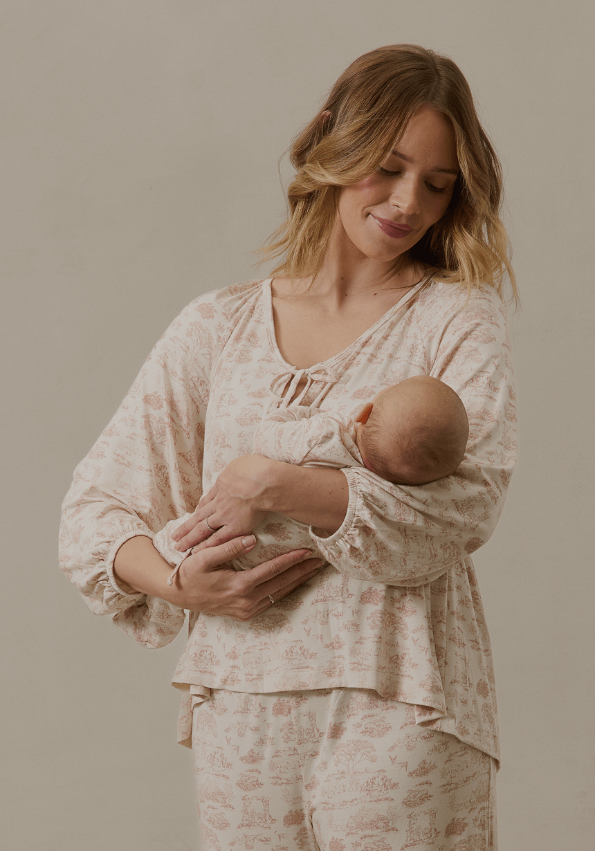 A mother cradles her newborn in her arms while wearing Solly Baby’s matching family sleepwear in a delicate toile print. The soft, breathable fabric drapes effortlessly, with gentle ruffle details adding a timeless charm. She gazes down at her baby with a serene smile, creating a moment of warmth and connection in effortlessly cozy loungewear.