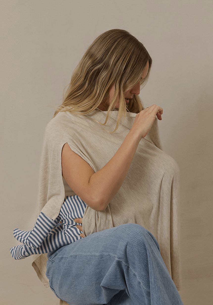 A mother with soft waves and a peaceful expression nurses her baby under a lightweight, breathable Solly Baby Nursing Cover in a soft oat hue. The draped fabric provides full coverage while allowing for easy eye contact and airflow. A tiny foot peeks out, clad in a striped sleeper, as she adjusts the cover for a cozy, intimate feeding moment.