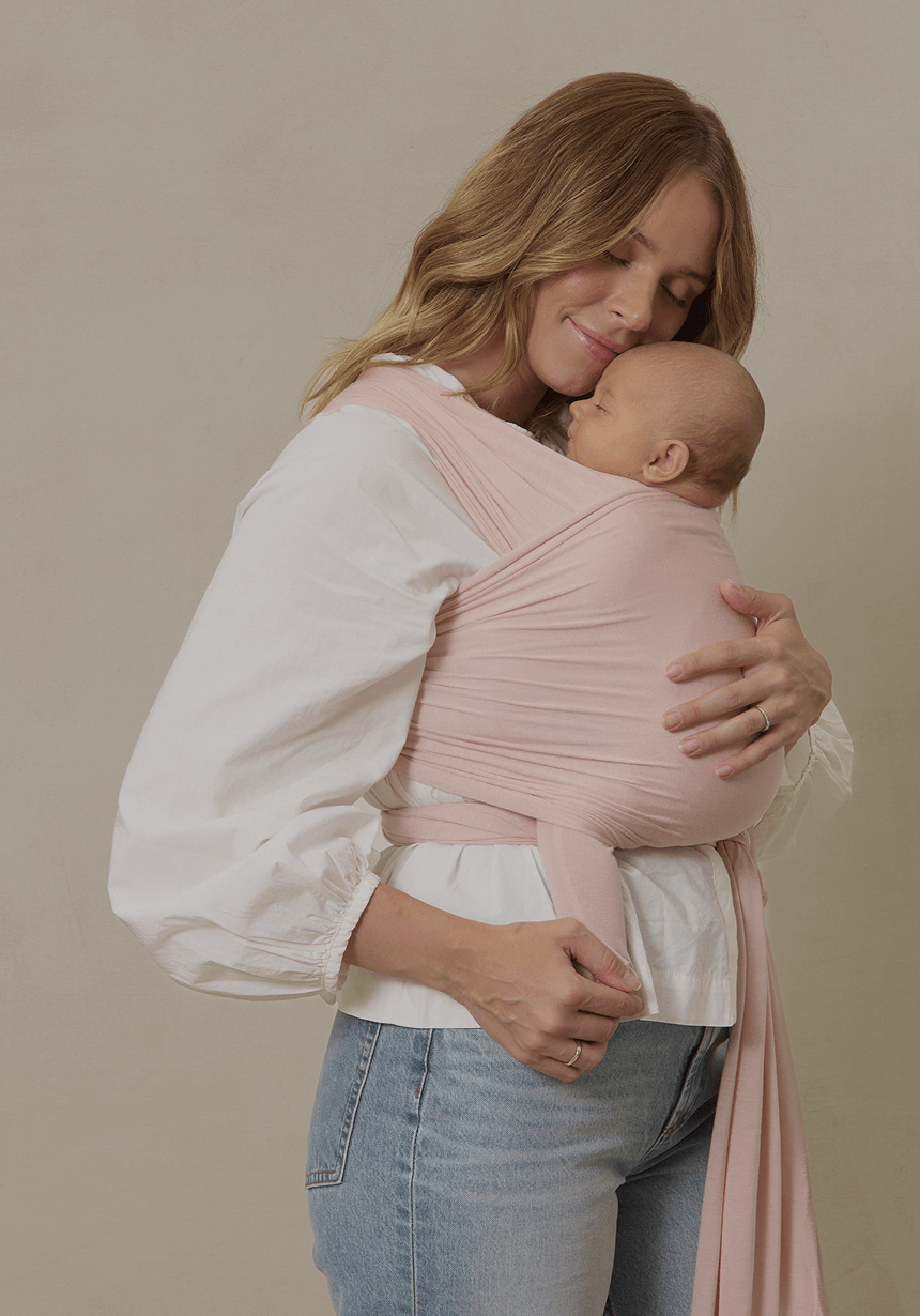 A mother with soft waves and a gentle smile cradles her newborn in a blush pink Solly Baby Wrap, creating a cozy, secure embrace. She wears a billowy white blouse and light-wash denim, standing against a neutral-toned background. The wrap’s lightweight, buttery-soft fabric molds perfectly to her and her baby, offering hands-free comfort and closeness.