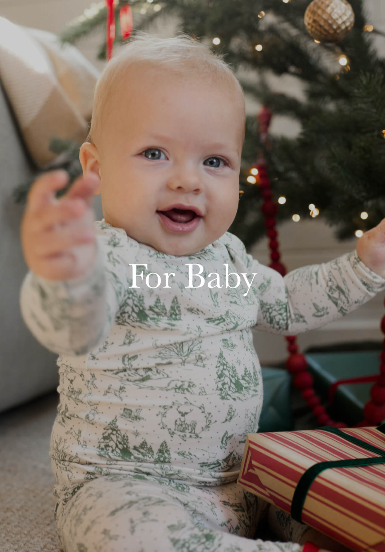 Baby smiling near a Christmas tree in a holiday-themed sleep set, holding hands up with a wrapped gift beside.