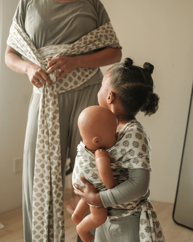 Woman putting on a green block pattern baby wrap while daughter looks up at her wearing her own matching play wrap holding a toy doll.