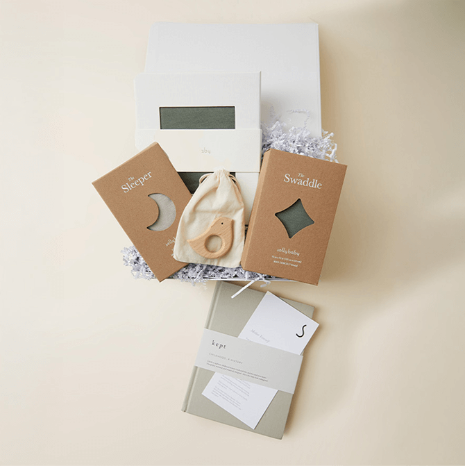 A beautifully curated gift box featuring Solly Baby essentials in a deep green hue. A soft sleeper and swaddle in kraft packaging, a matching wrap in its signature white box, a wooden bird teether in a cotton pouch, and a ‘Kept’ memory journal come together for a timeless newborn gift.