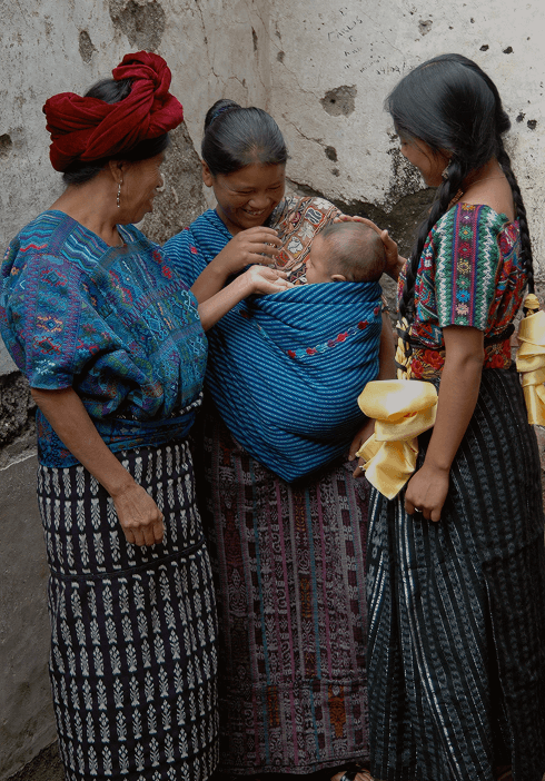 Three Guatemalan women sharing a moment with a baby nestled in a vibrant blue baby carrier. A celebration of tradition, culture, and community through Solly Baby's Ochoch partnership.