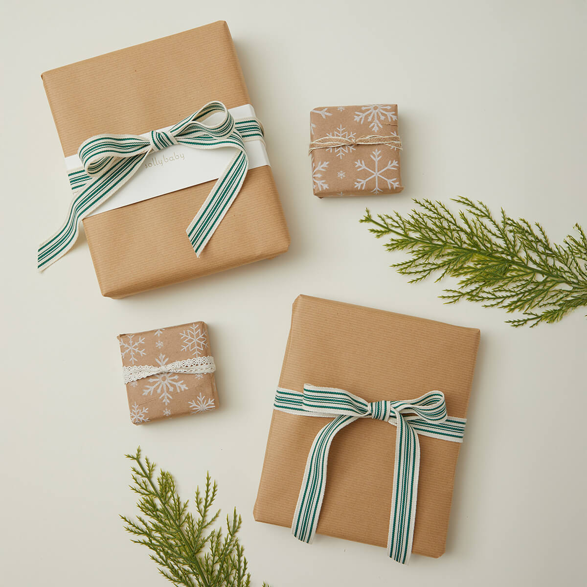 Flat lay of beautifully wrapped gifts featuring neutral kraft paper, one adorned with a green and white striped bow and another tied with delicate lace. Two smaller packages showcase snowflake-printed paper, creating a cozy and festive holiday aesthetic. Evergreen sprigs complete the seasonal, minimalist look.