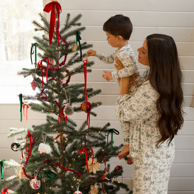 Mother holding baby, both in cozy pajamas, standing by a decorated tree.