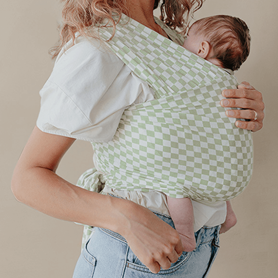 Mother wearing her baby in a Solly Baby Wrap featuring a sage green and ivory checkerboard print. Made from lightweight, breathable TENCEL™ Modal for a cozy, hands-free carry.