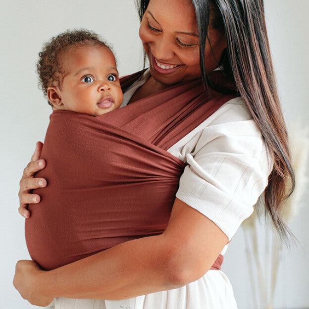 Smiling mother cradling her baby in a rhubarb-colored Solly Baby Wrap, with soft natural light in the background.