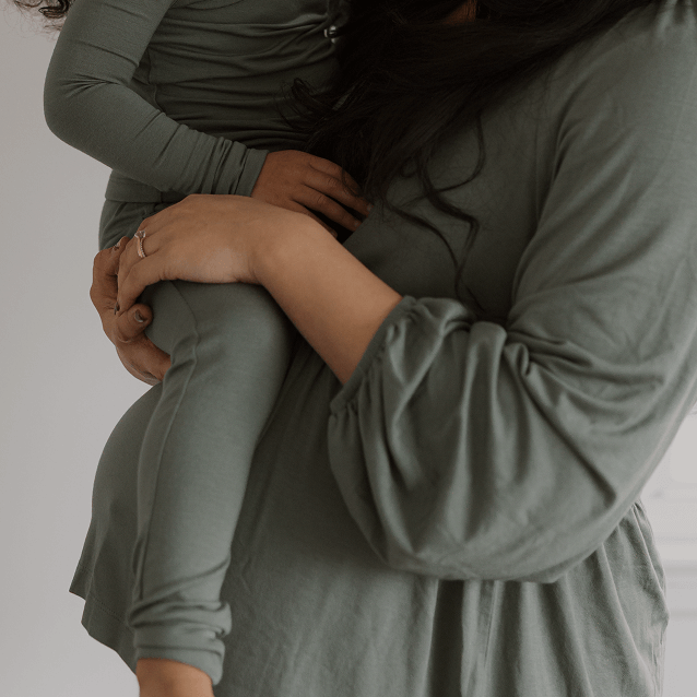 Close-up of a mother holding her child, both dressed in Solly Baby matching deep sage green loungewear. The mother’s hand gently cradles her child’s leg, showcasing the soft, breathable fabric designed for comfort and connection.