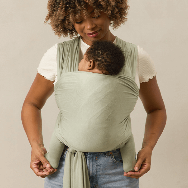 A mother stands holding her baby snugly in a Solly Baby Wrap in a muted sage green hue. The wrap is tied securely, providing a cozy and comfortable fit as the baby rests peacefully against her chest. She pairs the wrap with a soft, textured white tee and classic denim for a relaxed, effortless look.