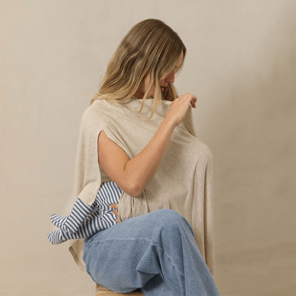 A mother sits comfortably while feeding her baby under a Solly Baby Nursing Cover. The lightweight, neutral-toned cover drapes effortlessly over her shoulders, providing full coverage and comfort while the baby's striped, knotted hat peeks out from underneath.