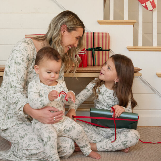 Mom and two children wearing matching green and cream winter toile pajamas, sitting by holiday gifts at the base of a decorated staircase.