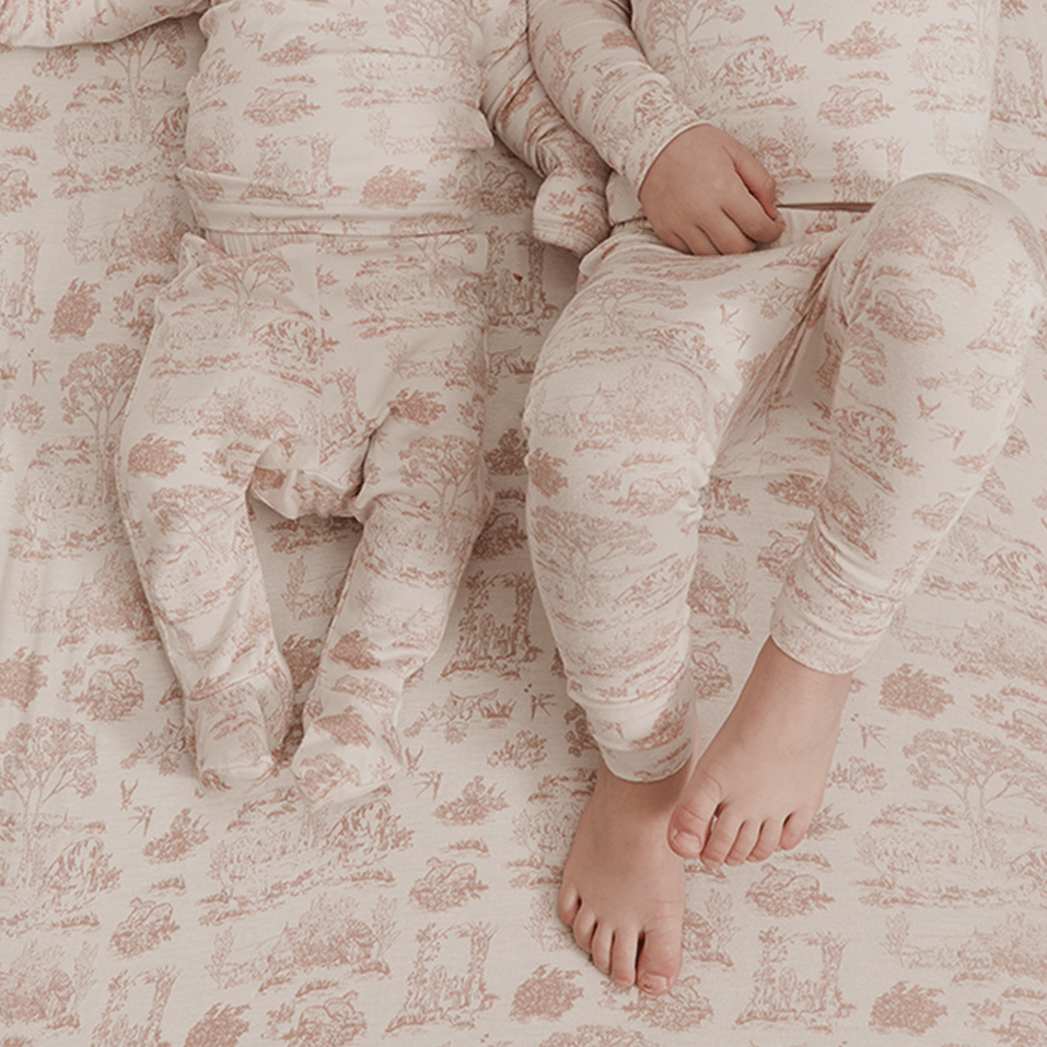 Close-up of baby and toddler wearing matching Solly Baby Sleep Sets in a rose toile print, resting on a coordinating crib sheet. Made from ultra-soft, breathable TENCEL™ Modal for all-night comfort.