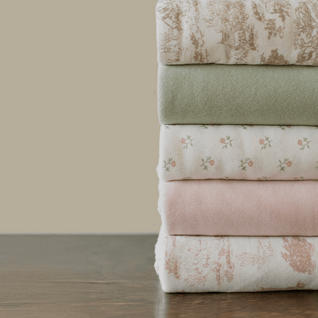 A neatly stacked collection of Solly Baby fabrics featuring soft pastel hues and delicate prints. The stack includes muted green, floral, and blush tones with elegant toile patterns, perfect for spring and Easter gifting.