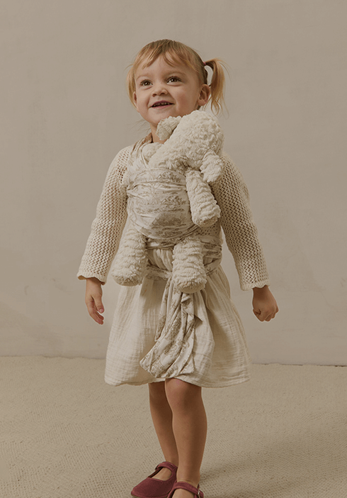 Toddler smiling while carrying a plush teddy bear wrapped in a miniature Solly Baby wrap featuring the delicate Toile design. Sweet bonding moments designed for little hands and big hearts.