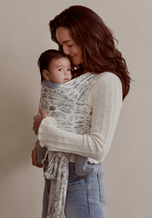 Alt text: Mother holding her baby close in a Solly Baby wrap featuring the delicate Toile design in soft cream and blue. A cozy, breathable carrier designed for comfort, connection, and hands-free bonding.