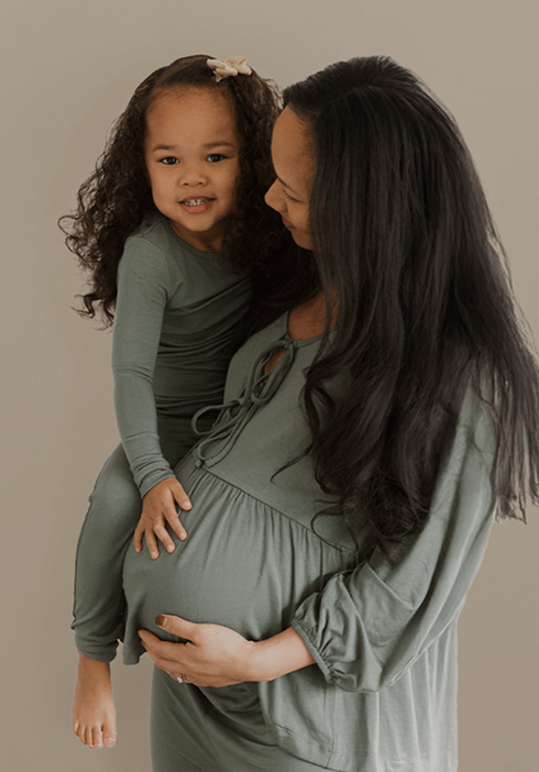 Mother and daughter wearing matching sage green Solly Baby sleepwear. The mother, visibly pregnant, cradles her belly while holding her smiling toddler dressed in a coordinating sage green sleeper. Soft, neutral tones create a cozy, family-focused moment.