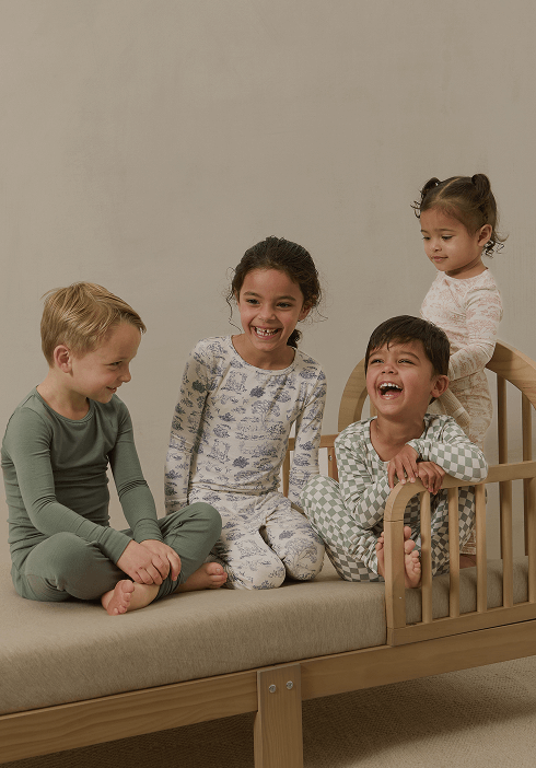 Three young children sitting on a wooden daybed, wearing Solly Baby pajamas in soft tones of sage green and Toile. Playful, relaxed, and designed for all-day comfort.