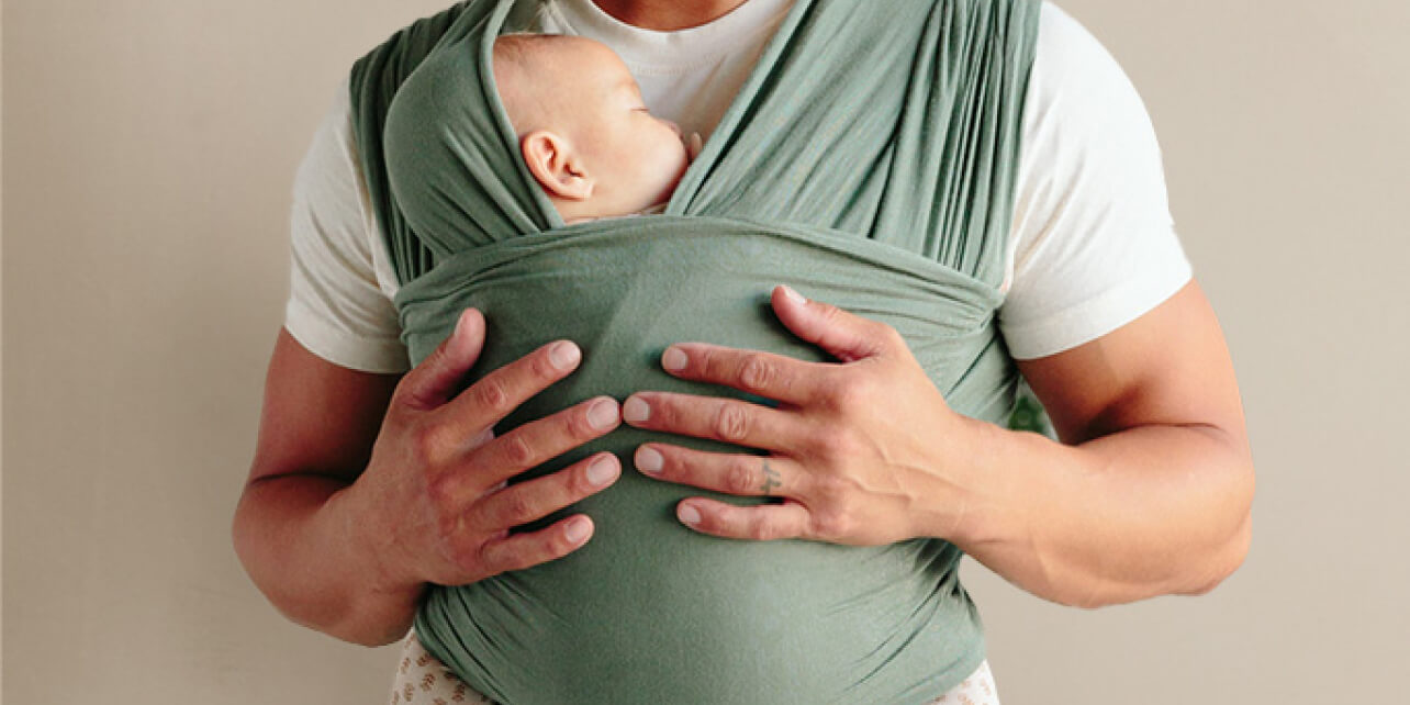 Father holding his baby snugly wrapped in a basil Solly Baby Wrap against a soft neutral background.