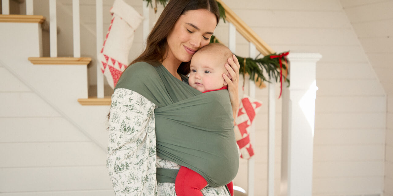 Mother carrying baby in a basil green wrap with a holiday-themed sleep set, standing in a decorated home for the season.