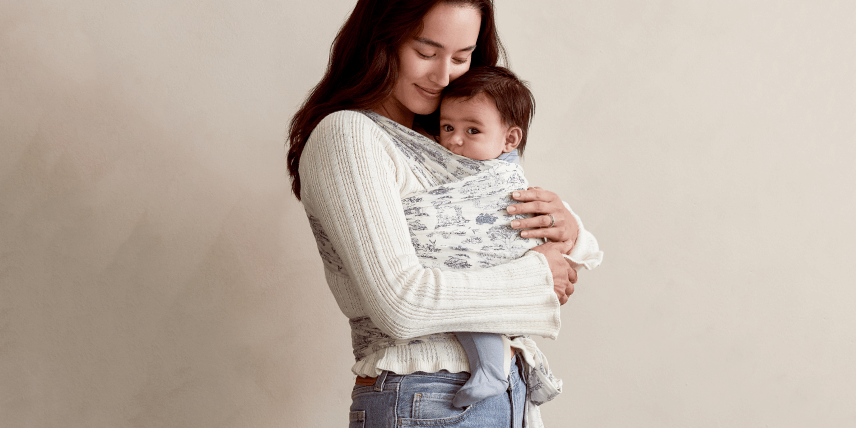 A mother smiles as she gently holds her baby close in a Solly Baby Wrap. The wrap features a delicate cream and soft bleu toile pattern, creating a serene and cozy look. The baby rests peacefully against her chest while she stands in a soft cream knit sweater and relaxed denim, embodying warmth and connection.