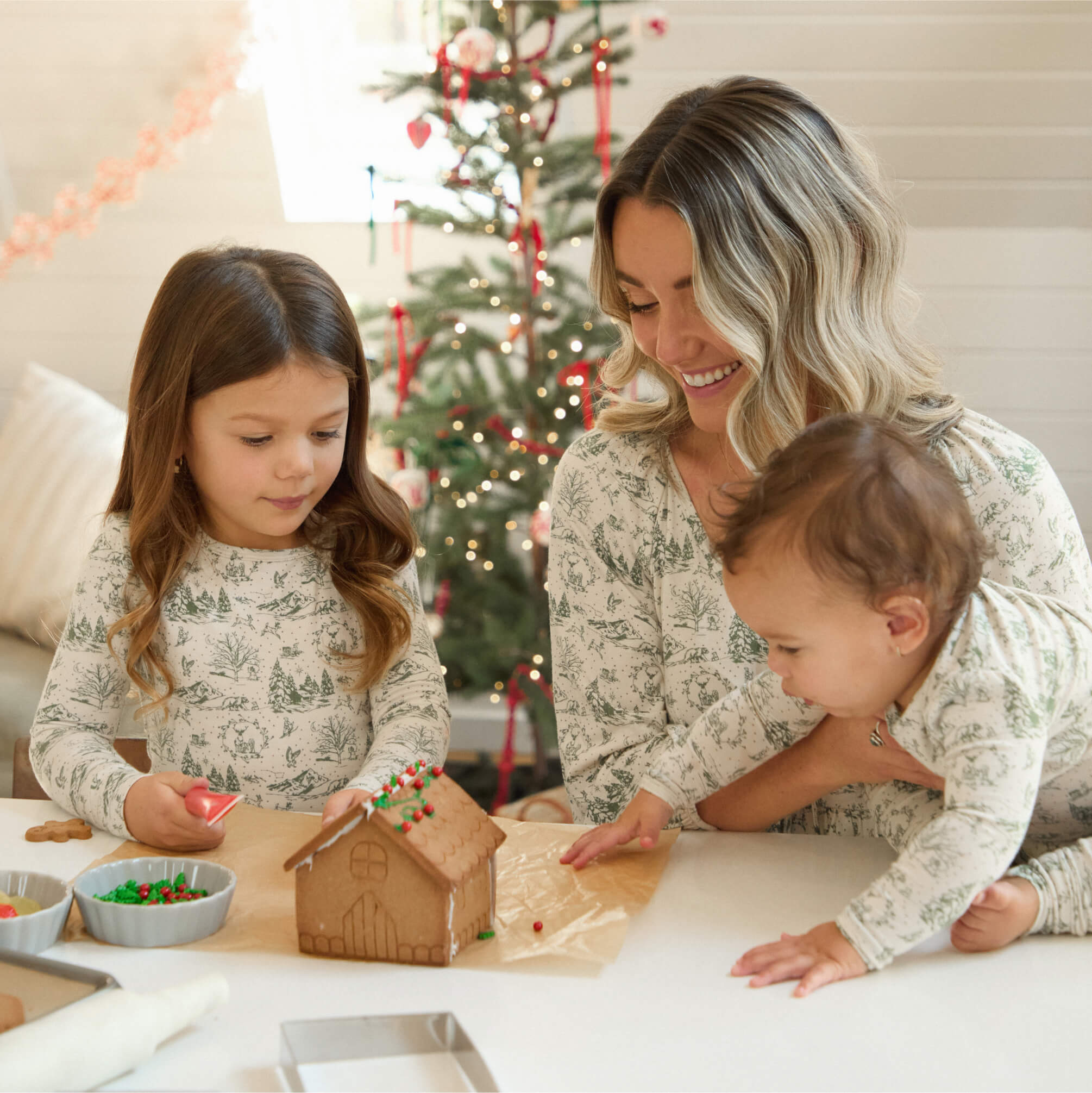 Family gathered around a table decorating a gingerbread house, with holiday lights on the tree behind them.