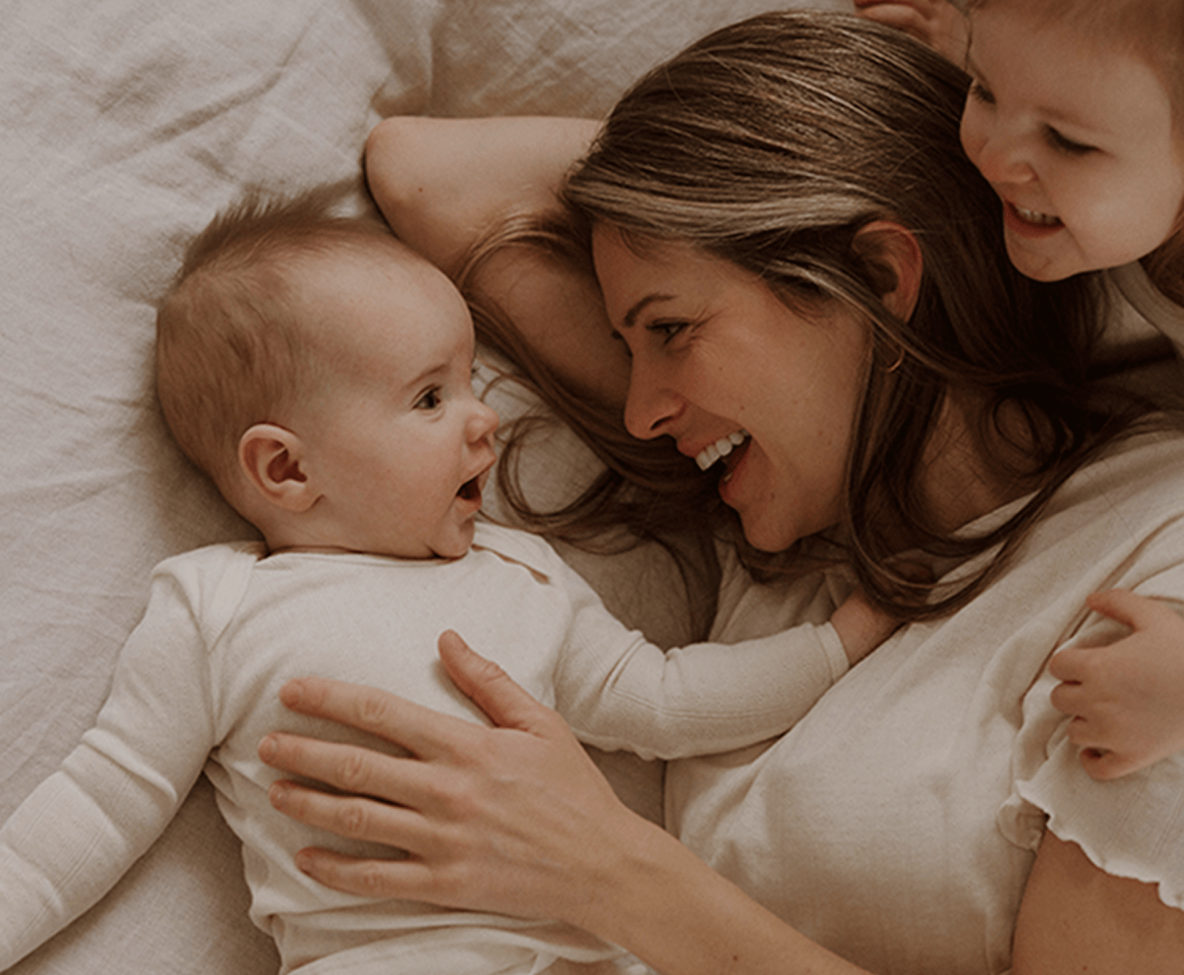 A warm, cozy lifestyle shot of a woman and two babies lying on a bed, all wearing matching cream-colored long-sleeve baby bodysuits. The scene is bathed in soft, warm, golden-hour light.