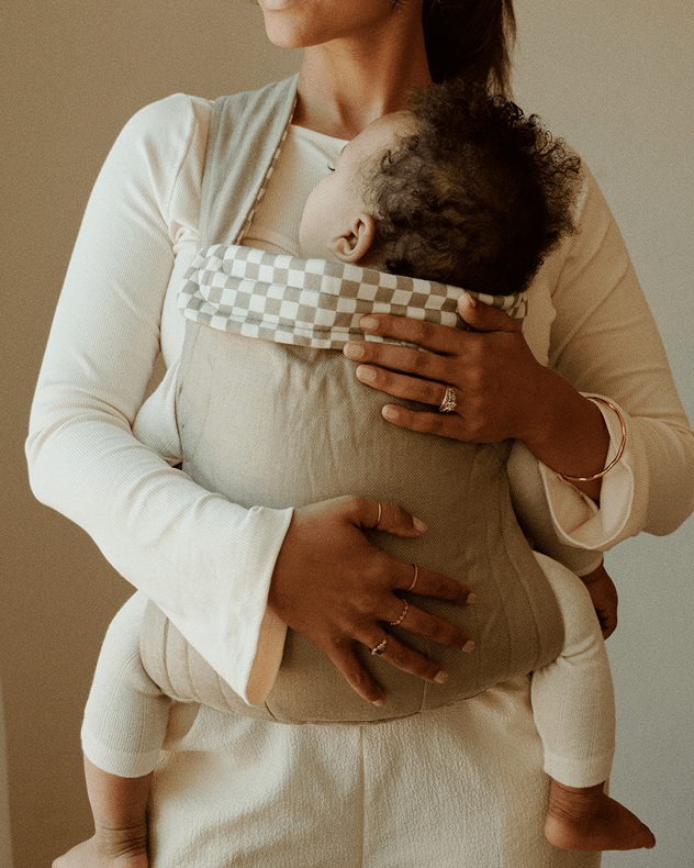 A woman wearing a cream-colored long-sleeve baby carrier with a baby inside. The carrier has a green body and a checkered patterned waist strap in beige and white.